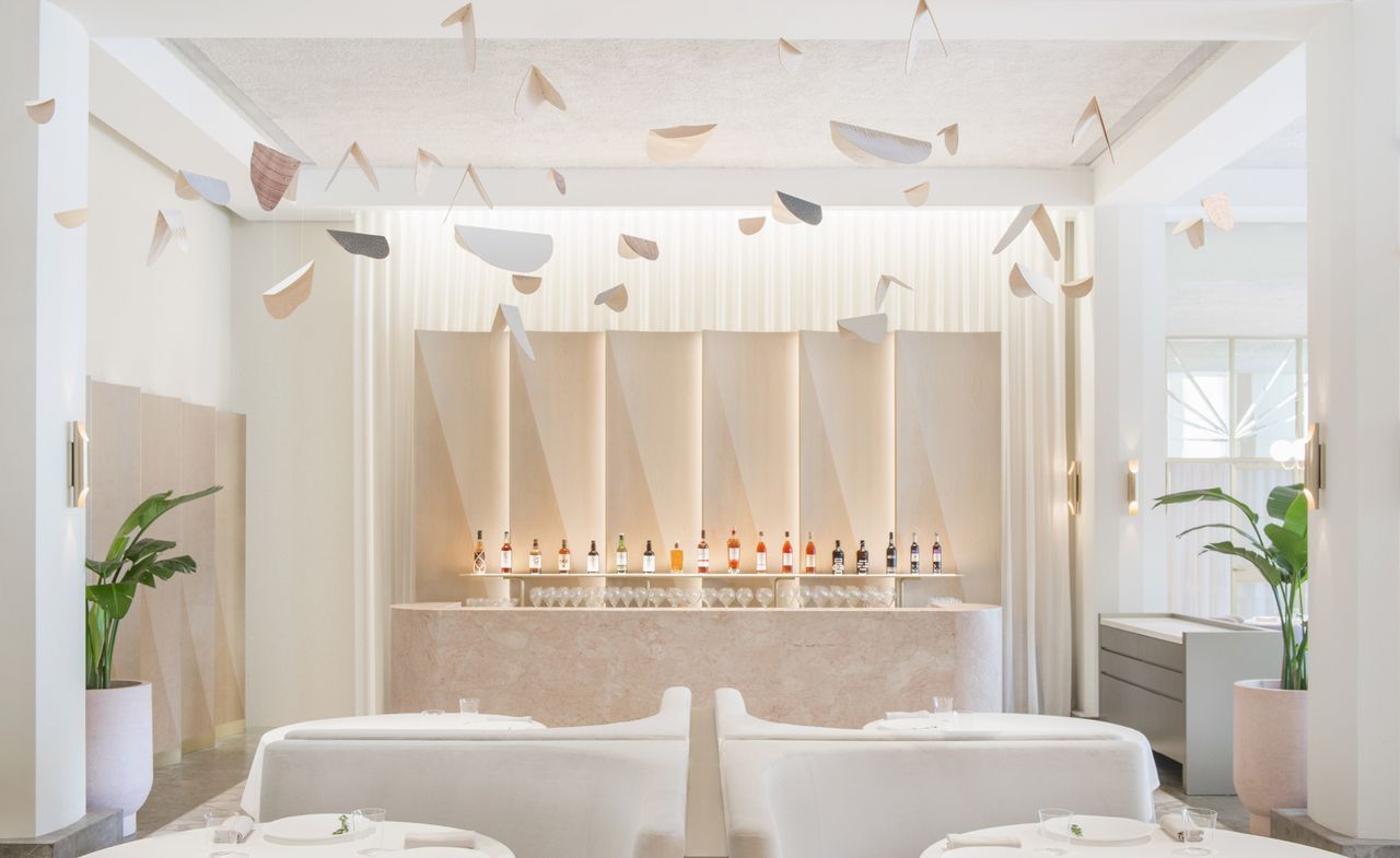 Interior view of the bar and dining area at Odette featuring light coloured decor, tables with tableware, chairs, green plants in white pots, shelving behind the bar with bottles of drinks and glasses and shapes hanging from the ceiling that resemble butterflies