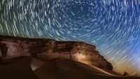 Star trails circle above a desert at night as meteors streak through the sky.