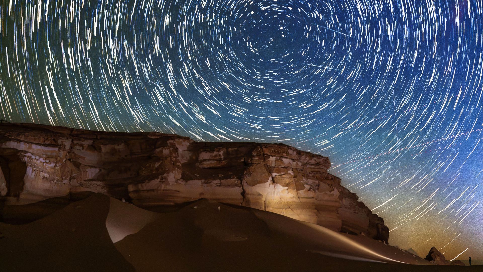 Star trails circle above a desert at night as meteors streak through the sky.