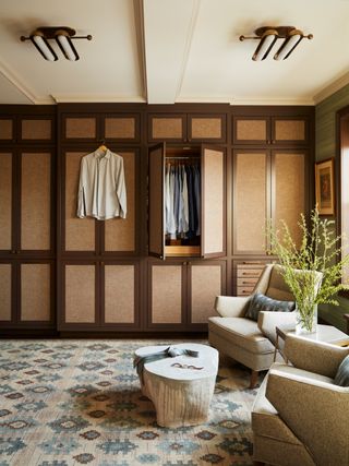 dressing room with wall of closet doors, patterned rug, footstool, two armchairs, side table, shirt hanging up, 2 doors open, retro ceiling lights