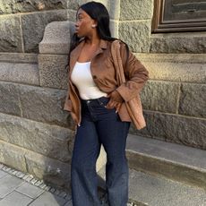 A woman wears a brown suede jacket, white tank top, jeans and adidas sneakers.