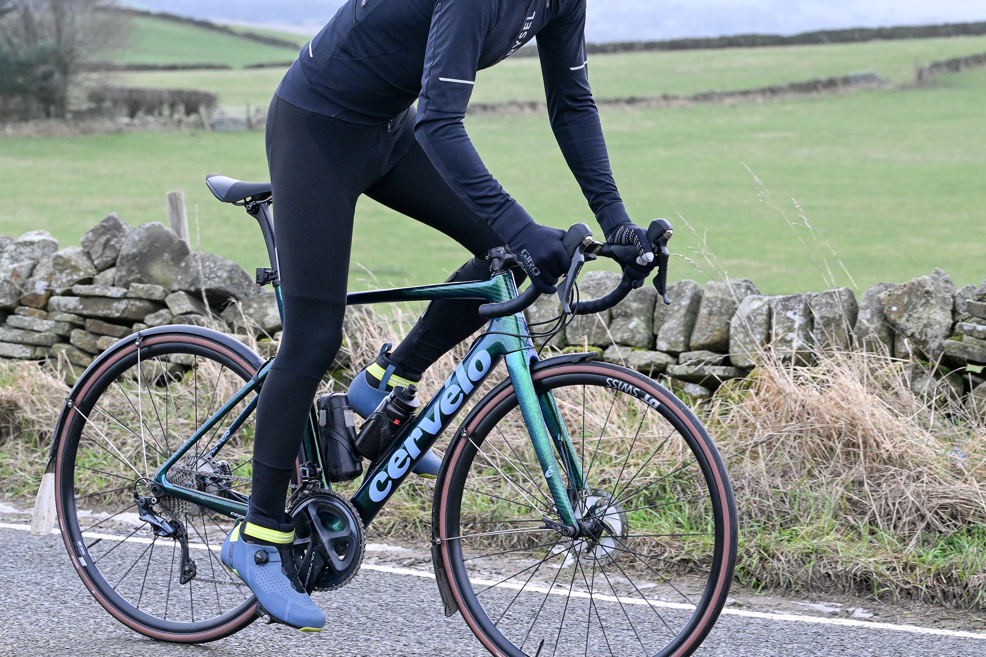 side view of a man riding a green Cervelo road bike wearing black tights, black jacket and blue shoes