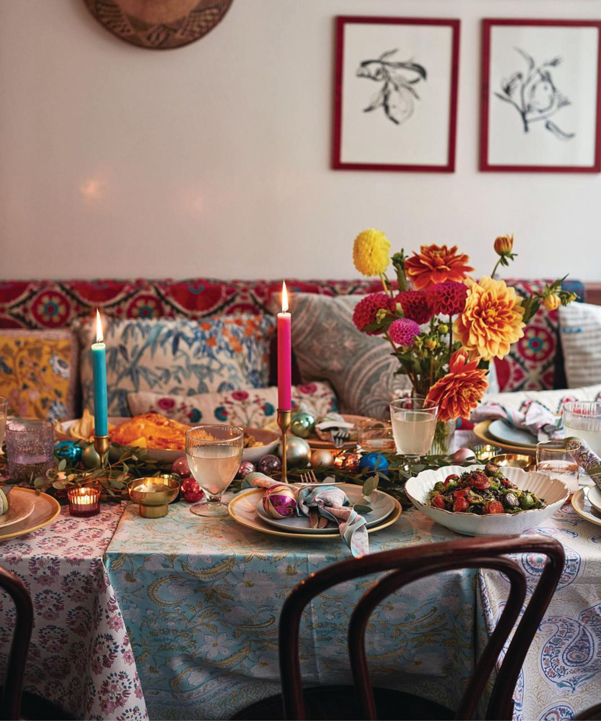 A festive Christmas dinner table with colorful layered and patterned tablecloths, bright taper candles in gold holders, a vase of orange, yellow and pink flowers, brown wooden chairs, and assorted dishes. The walls are white and on the background wall there are two botanical prints in red frames.