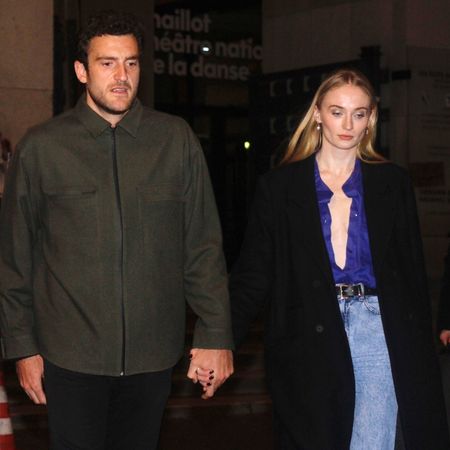 Sophie Turner wearing an open blue button down with a black wool coat and light wash jeans 