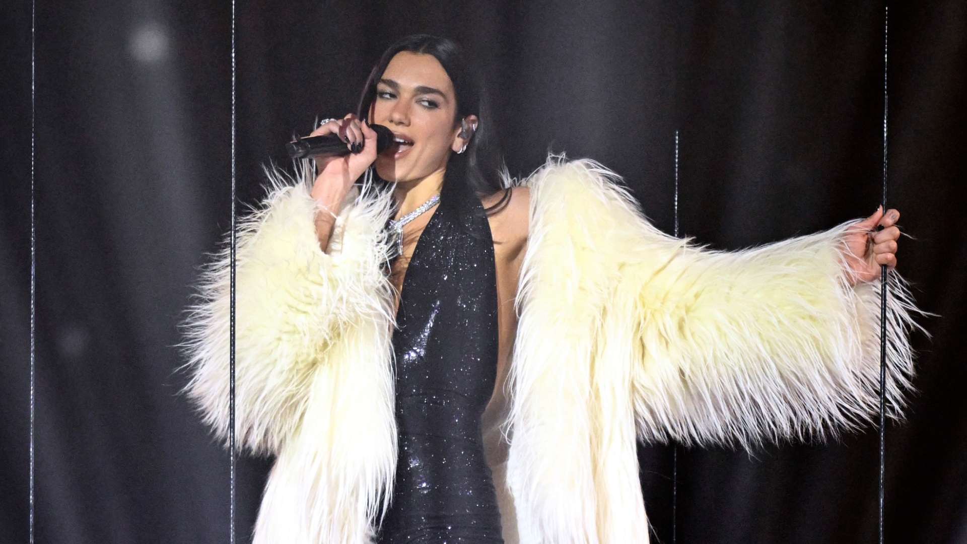 Dua Lipa performs at the 2026 BRIT Awards wearing a sparkly black plunging body suit, a white fur coat, and knee-high boots with fishnets