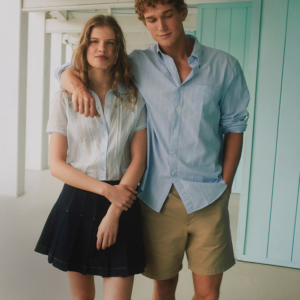 A man in a blue button up shirt and tan shorts with his arm around a woman in a light blue button up short sleeved shirt and black skirt
