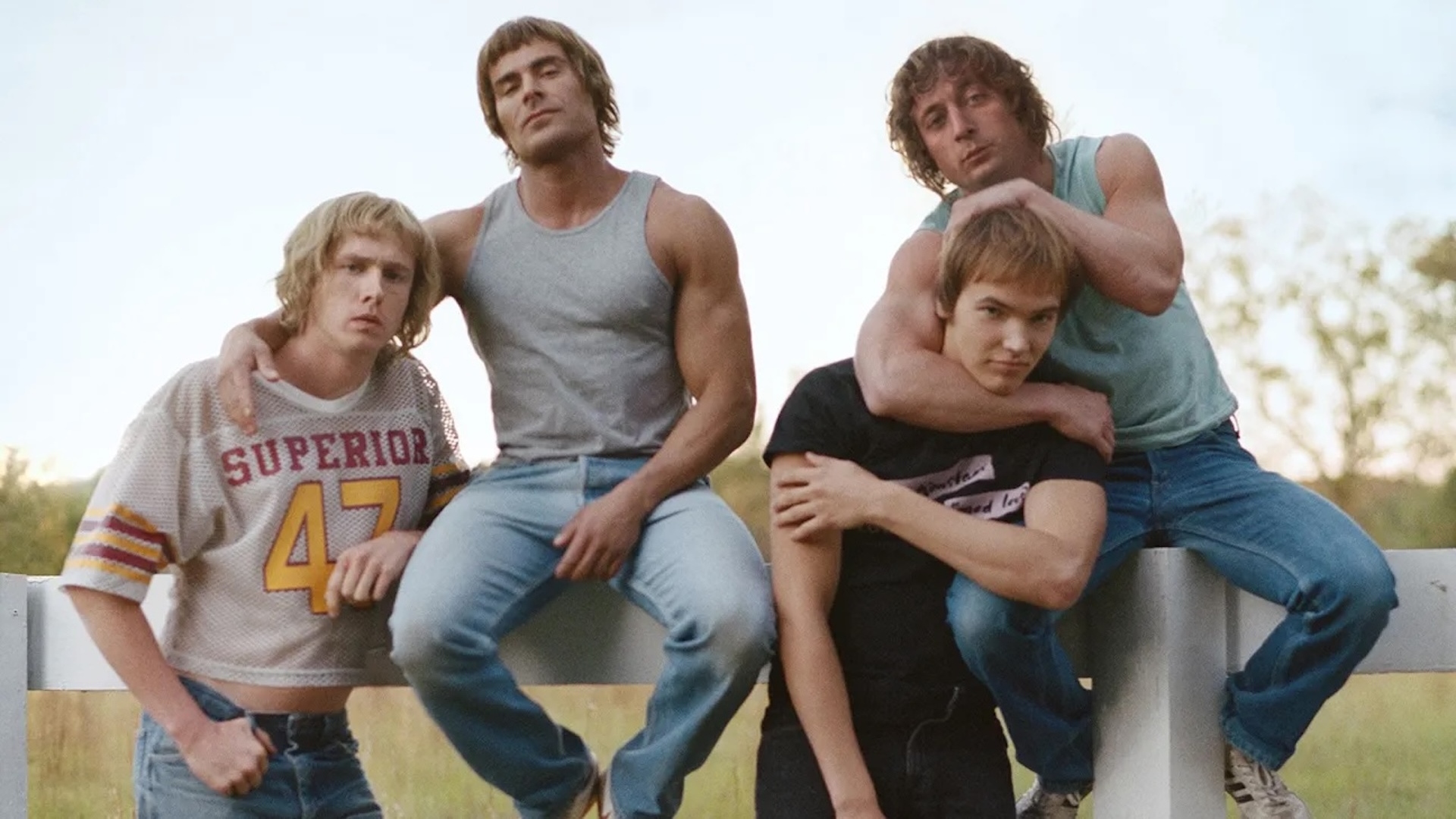 Four wrestlers from the film The Iron Claw sat on a fence 