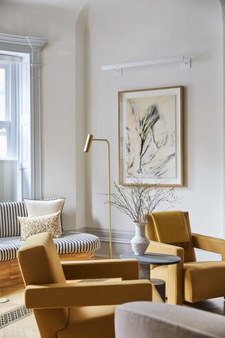 neutral living room with two mustard armchairs, a brass floor lamp, artwork, and striped window seat