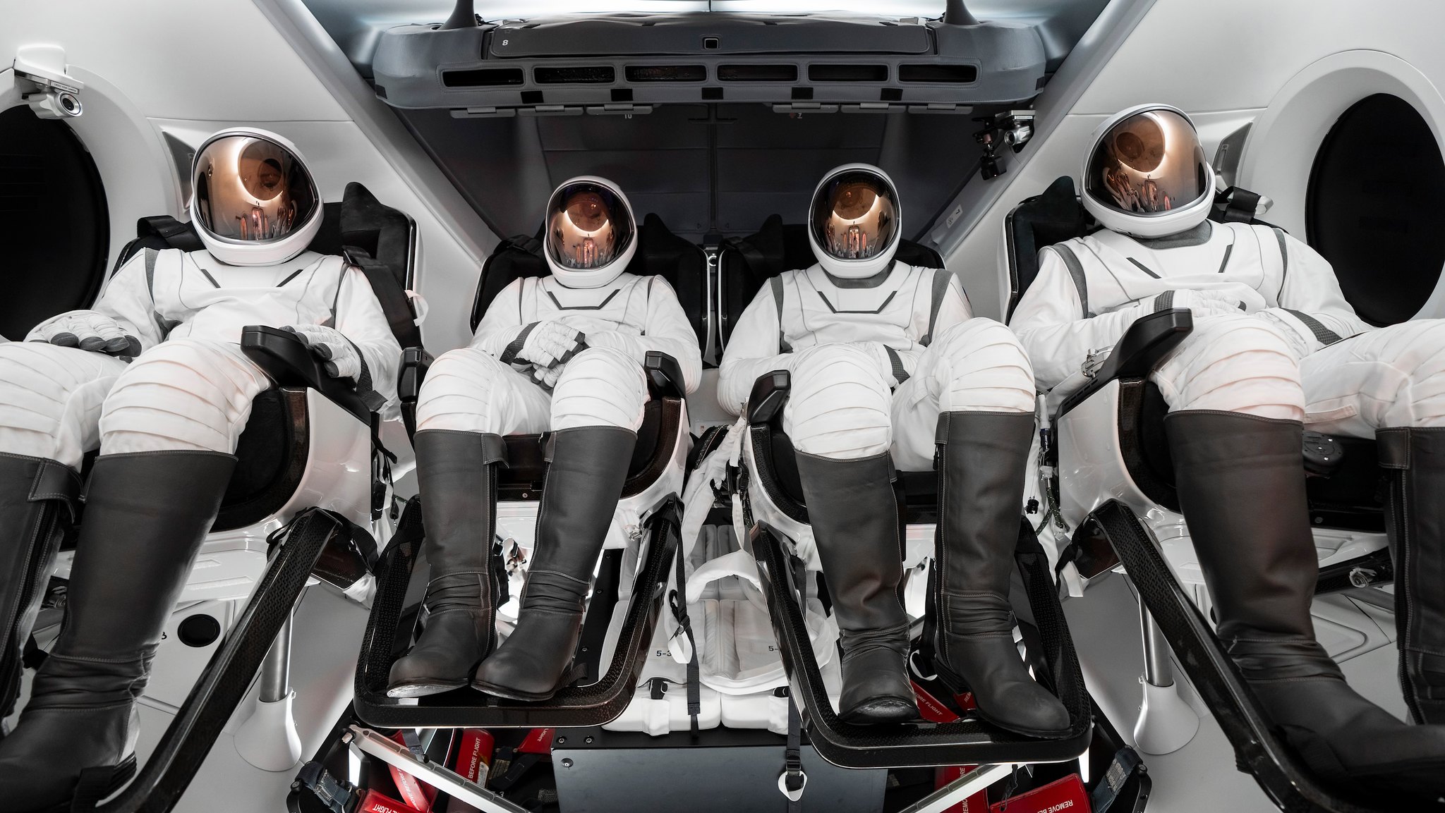 Four people in white spacesuits with gold shields sit inside the cabin of a white spacecraft.