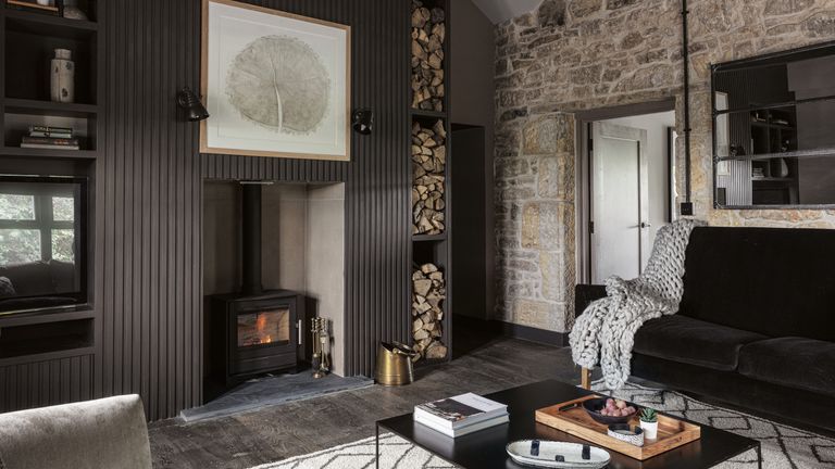 Living room with original brick wall, large fireplace with woodburning stove surrounded by dark painted slatted panelling. Black velvet sofa and sleek black square coffee table