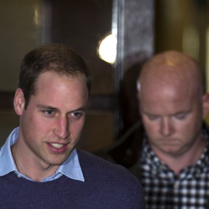 Prince William wearing a blue sweater with two men walking behind him