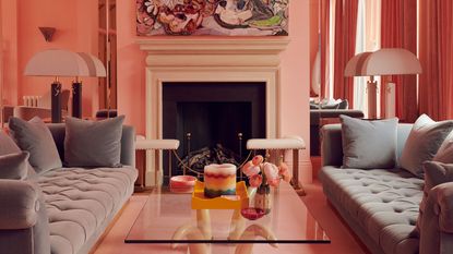 A modern pink living room with two gray sofas facing each other with a square glass coffee table in the center in front of a fireplace with artwork above it and large white table lamps either side