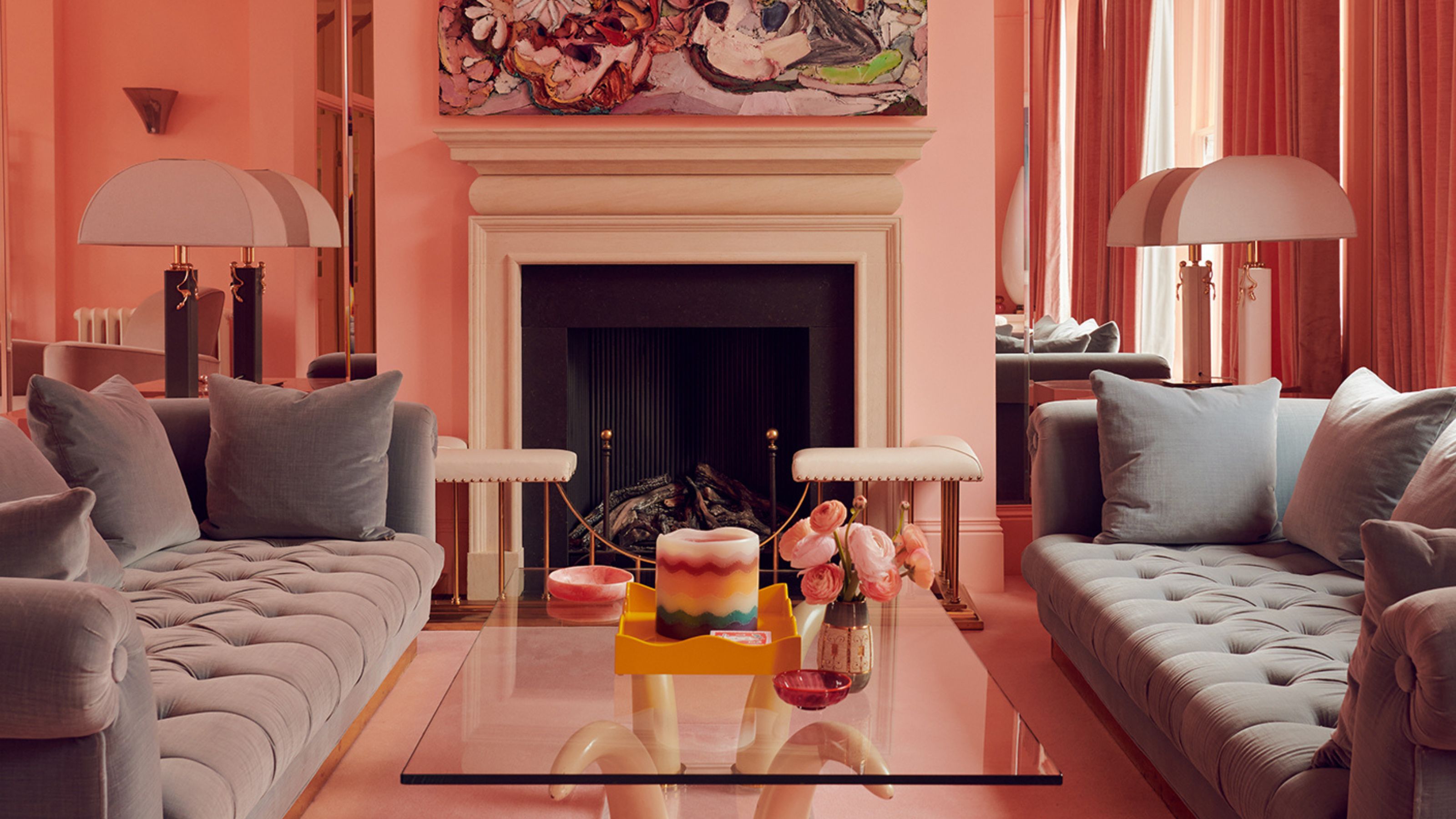 A modern pink living room with two gray sofas facing each other with a square glass coffee table in the center in front of a fireplace with artwork above it and large white table lamps either side