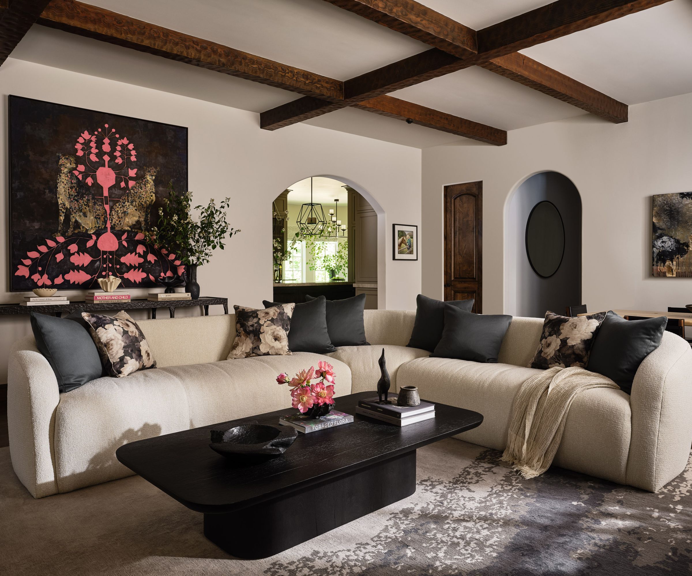 Cozy living room with a large L-Shaped cream sofa with black and floral pillows, an oval-shaped black wood coffee table, a patterned rug, white walls with arch doorways, and wooden beams