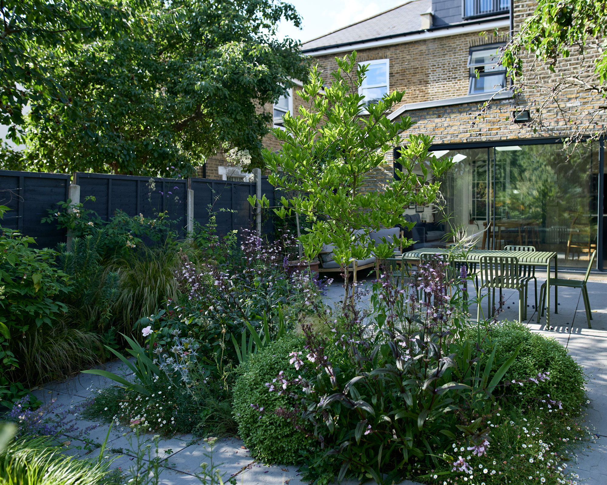 shade garden design with repeat planting of foxglove beardtongue, hebe and sea holly in flower bed, path, outdoor furniture set on patio, black fence and pleached trees