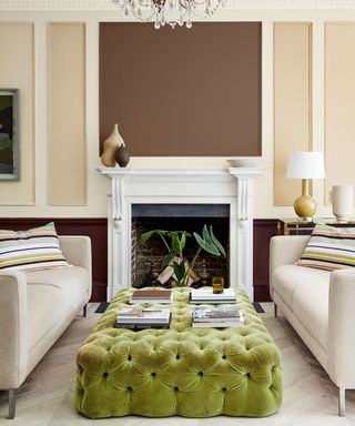 Brown and cream painted living room with whit paneling, white fireplace, two cream sofas facing each other, bright green tufted ottoman placed between two sofas, light gray wooden flooring