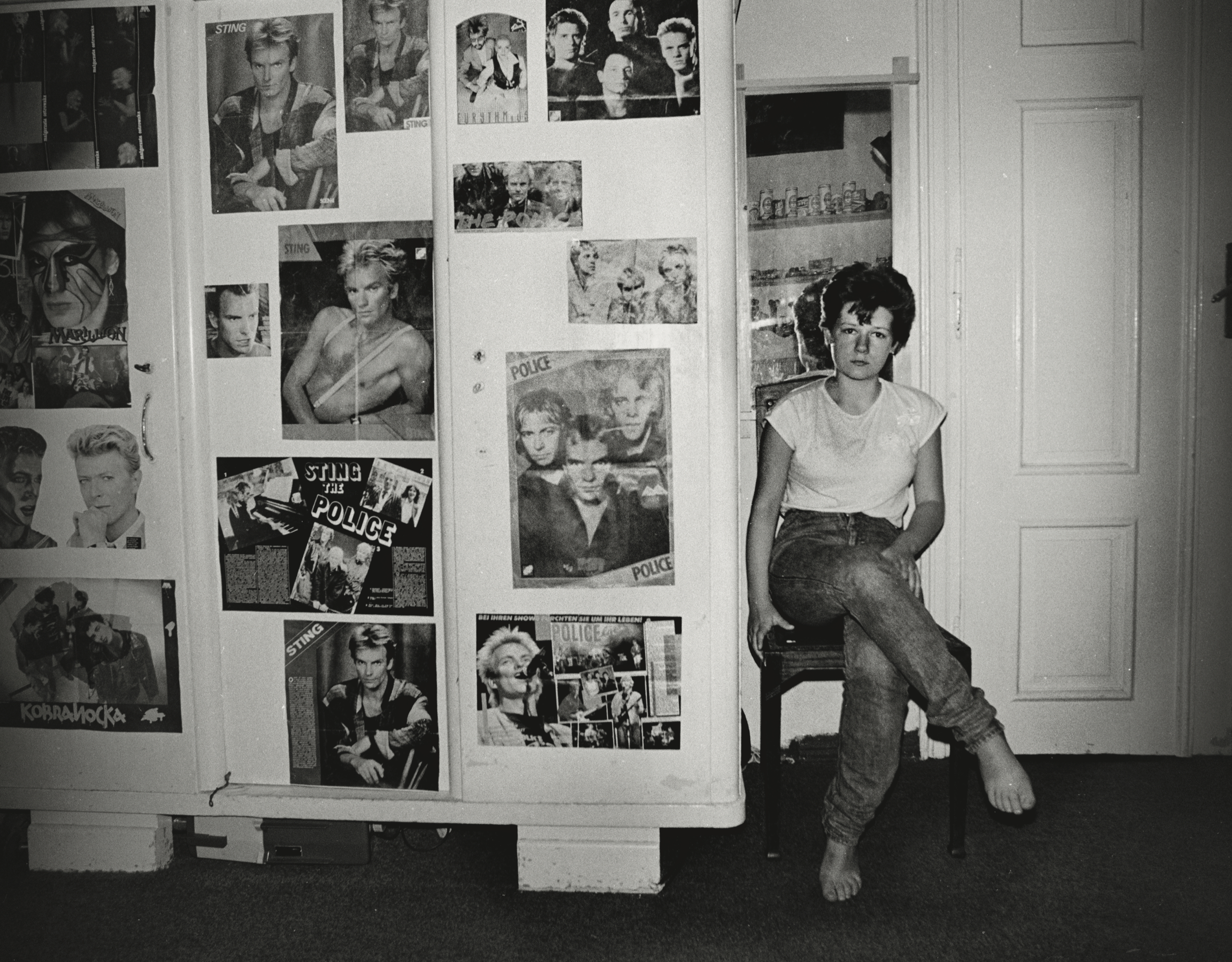 Black and white photograph of a young woman sitting on a chair next to a wardrobe decorated with several posters of musician Sting, and rock band ‘The Police.’