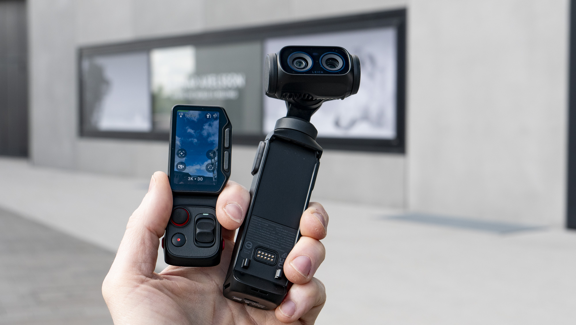 Hand holding the Insta360 Luna Ultra vlogging camera and its screen, outdoors in a grey urban scene