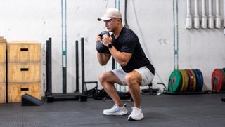 Man in gym performing goblet squat holding kettlebell. His knees are bent, his hips are pushed back and the kettlebell is held just below his chin in both hands. The model is wearing a blue T-shirt and white baseball cap, shorts and trainers.