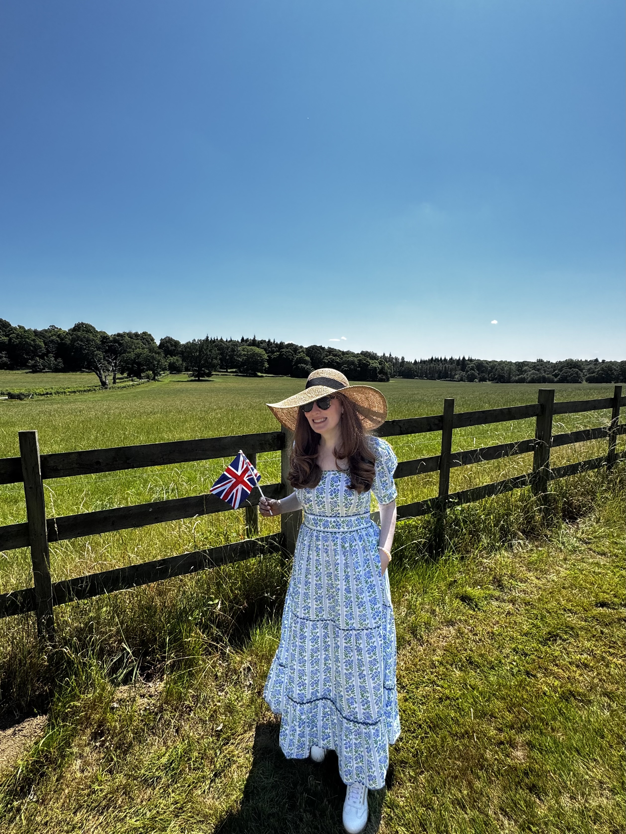 Kristin Contino wearing a blue floral dress and sun hat standing against a fence in the English countryside