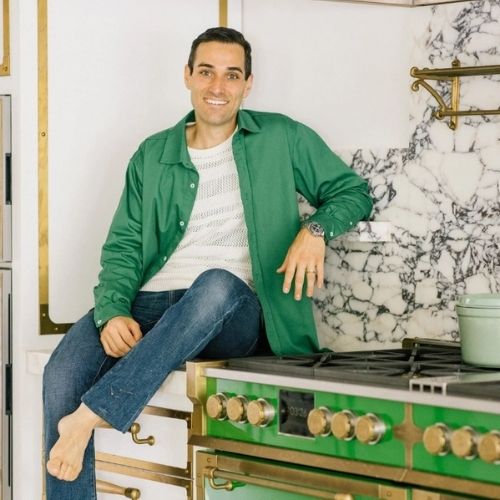 Image of a man in a white top, jeans, and a green button-down sitting on the countertop of a kitchen.