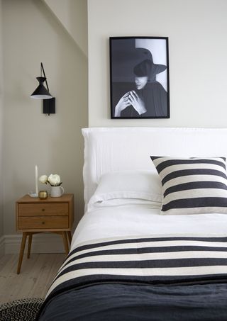 black and white bedroom with black wall light, black and white artwork, white bedding with black and white stripe cushion and blanket, mid-century modern nightstand, wooden floorboards