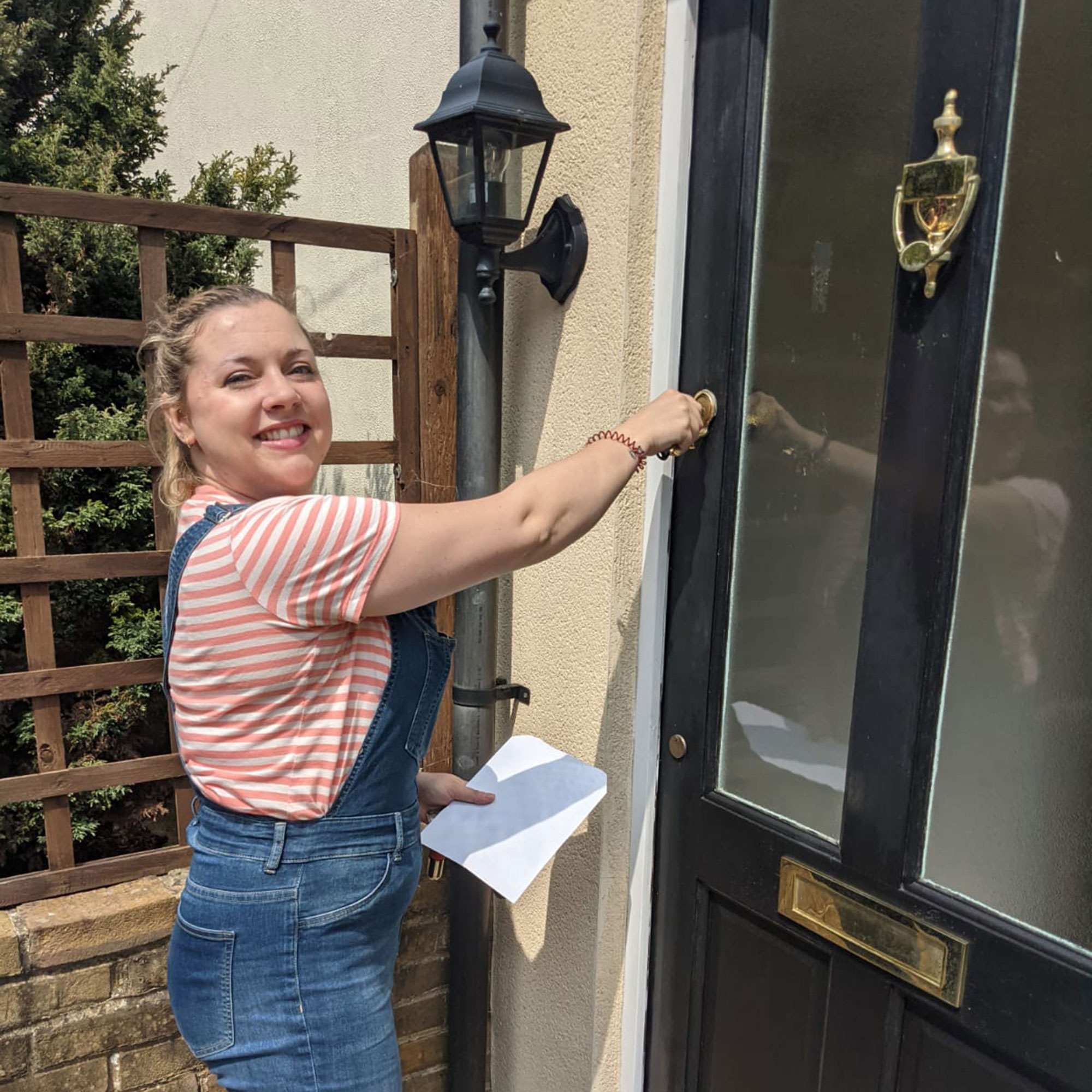 White blonde woman in dungarees in front of a door