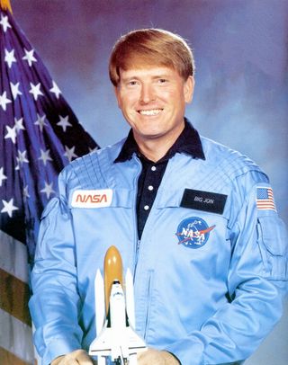 a smiling man in a blue jacket holds a model of the space shuttle while standing in front of an american flag