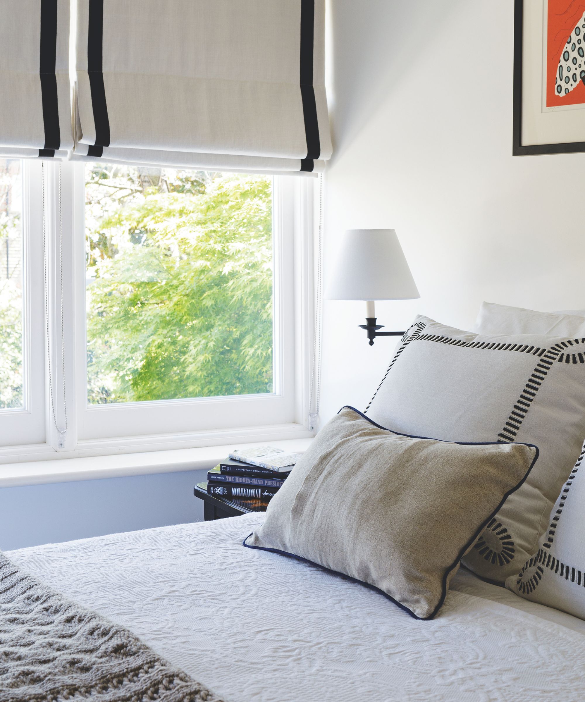 A white bedroom with a bed with white bedding in the foreground, with a grey cushion at the foot and two large square white and black cushions at the head, with a rectangular neutral beige cushion leaning up against them. To the left of the bed is a white wall lamp, and the corner of a bedside table with a stack of books on it is seen. Behind is a large window with green leaves seen beyond, and a cream and black blind.