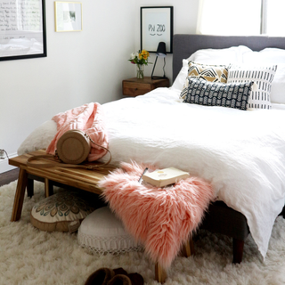White bedding on bed, with wooden bench at foot end layered with coral fluffy faux fur layer, with round boho floor cushions beneath, and mud cloth inspired scatter pillows on bed.