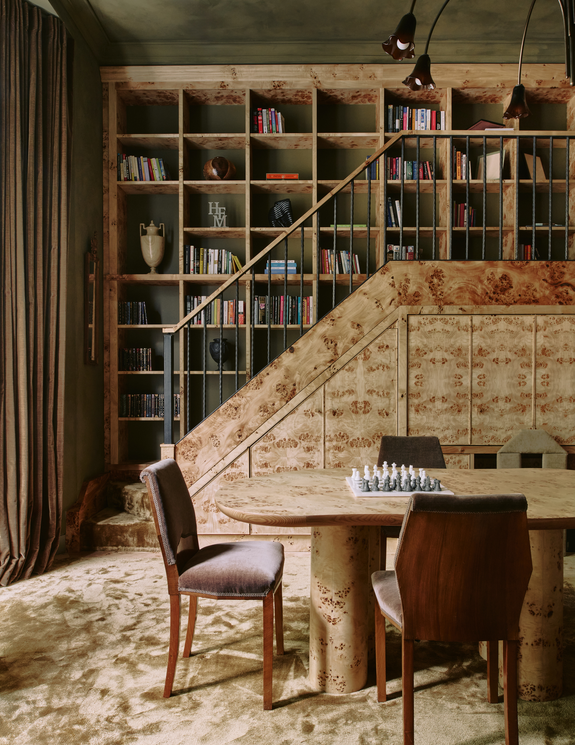 Image of a library room with limewashed olive green walls and burl wood cabinetry, built-ins, and games table. There is beige carpet in the room and a chess board on the table.