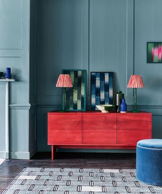Blue living room with red sideboard, two red table lamps, colorful artwork