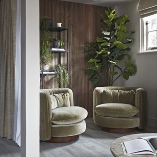 A cosy seating nook with fluted wood walls, green velvet chairs and house plants with natural light streaming in from a window