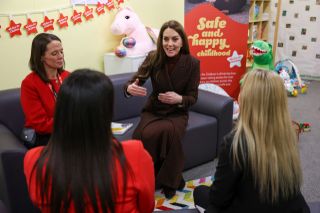 Kate Middleton sitting on a sofa at a prison nursery wearing a burgundy plaid coat and talking with. her hands while surrounded by 3 women and toys on the floor