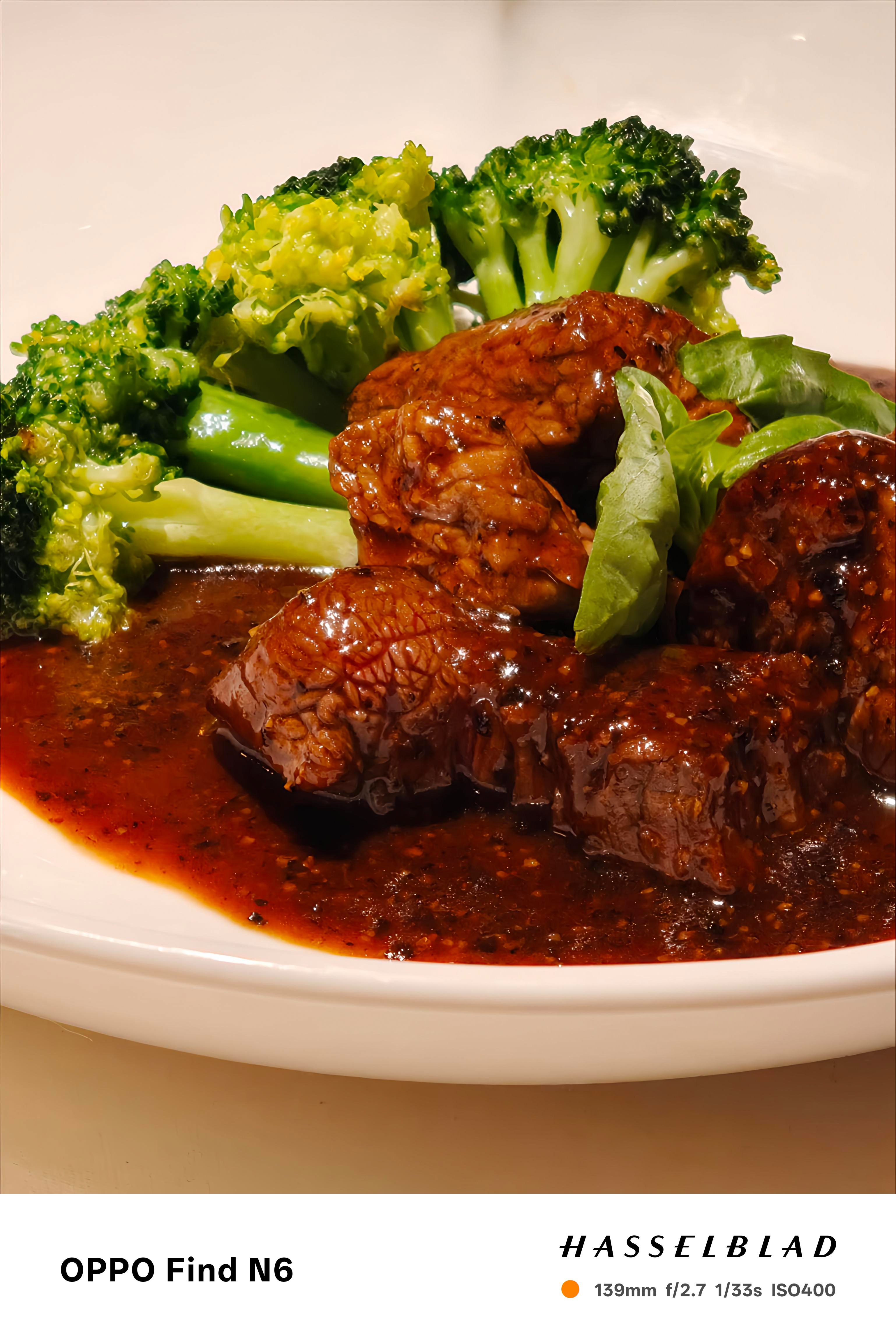 A food photography shot showing tender pieces of beef glazed in a dark, savory pepper sauce, served alongside bright green steamed broccoli florets on a white plate.