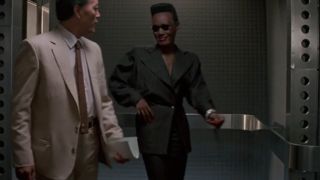 Grace Jones wears a deadly smile in a black suit as she escorts someone through a room in A View To A Kill.