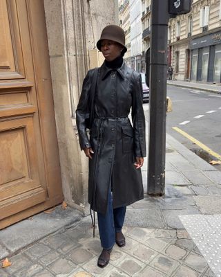 Woman wearing dark jeans, black leather coat, and brown hat standing on Paris street.