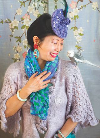 A photo of Terri Wong in an over 60 outfit featuring a lilac knit top, blue and green scarf, and a purple feathered headdress with a cockatiel bird on her shoulder.