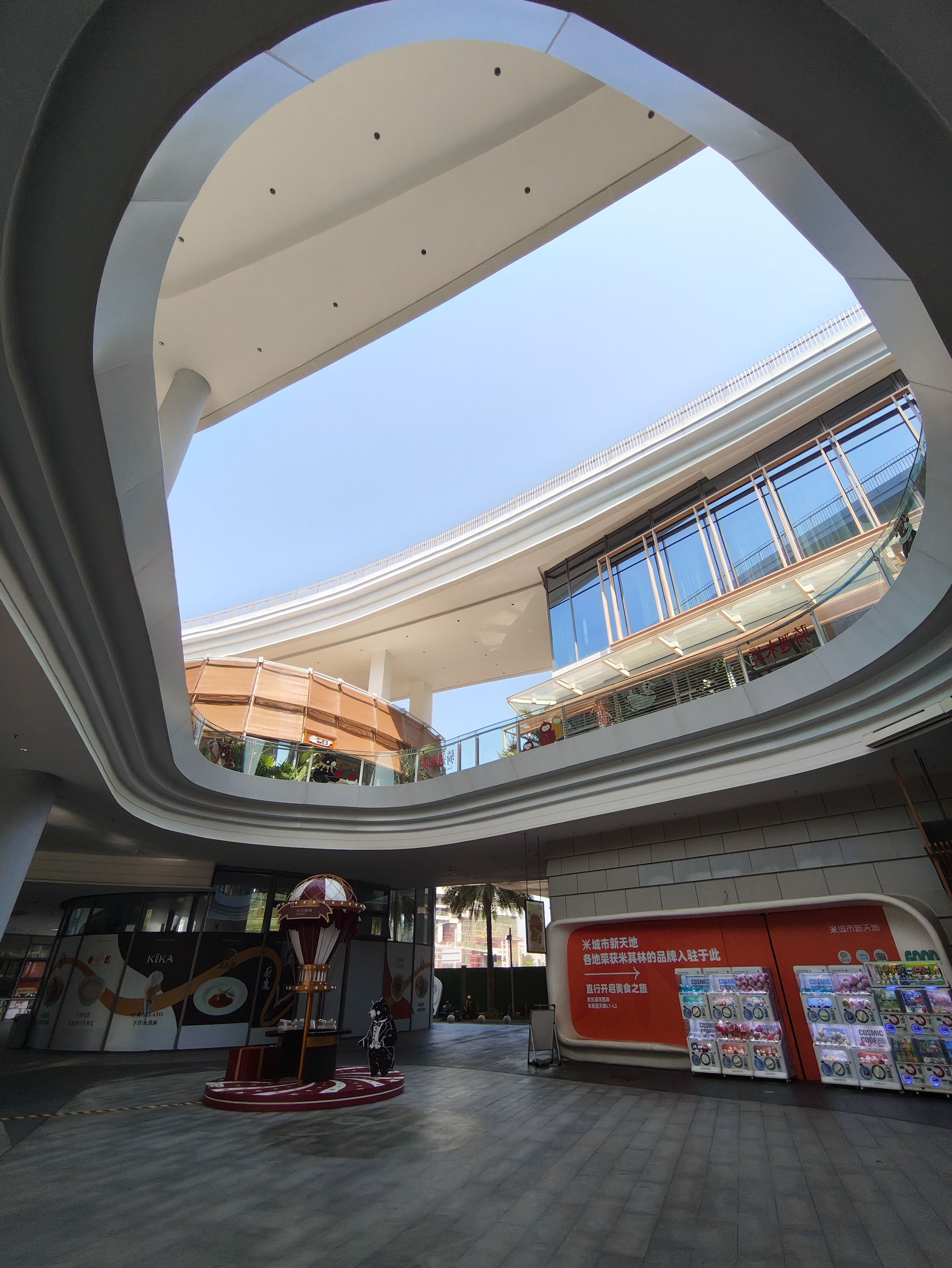 A wide-angle, low-angle shot looking up at the interior courtyard of a modern, multi-level shopping complex. The building features smooth, white, flowing architectural curves that frame a bright blue sky. On the ground floor, there is a small decorative hot air balloon display and a row of colorful capsule toy vending machines against an orange wall.