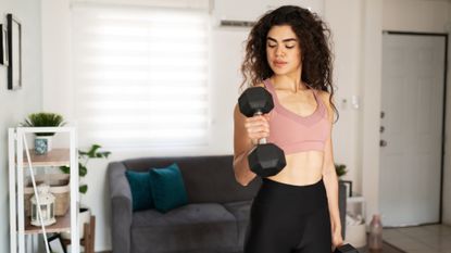 woman curling a dumbbell up to her biceps standing in a living room setting. she's wearing a light pink crop top and dark bottoms.