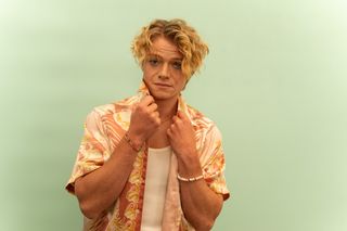 Ricky September (Tom Rhys Harries), who is tanned and has wavy dirty blond hair, poses with his hands clasping the collar of his short sleeved Hawaiian shirt