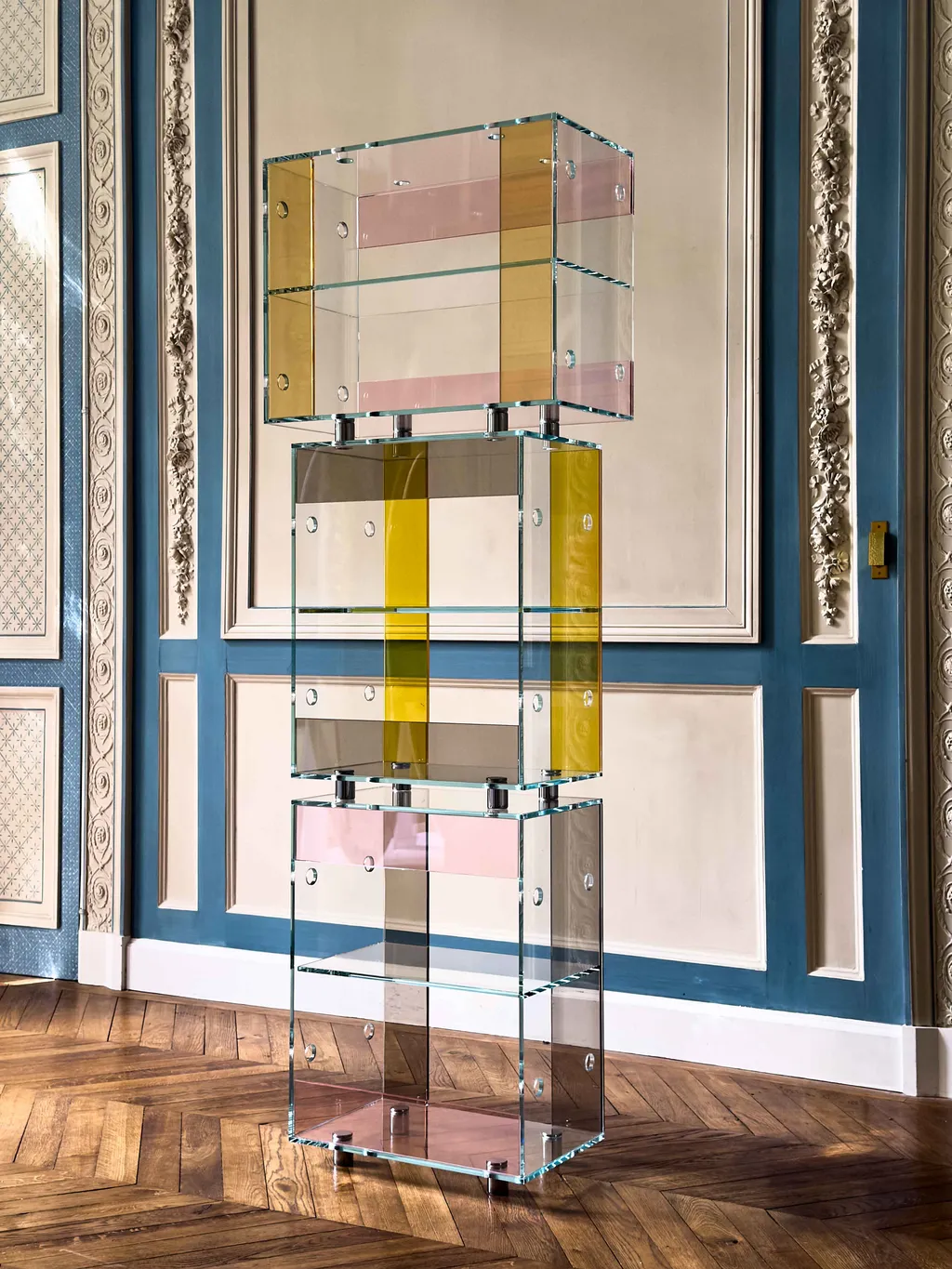 A towering glass sculpture made of transparent pink and yellow-tinted panels bound together into a three-part totem sits in a wood-paneled room with blue and white, stuccoed walls and wooden floors.