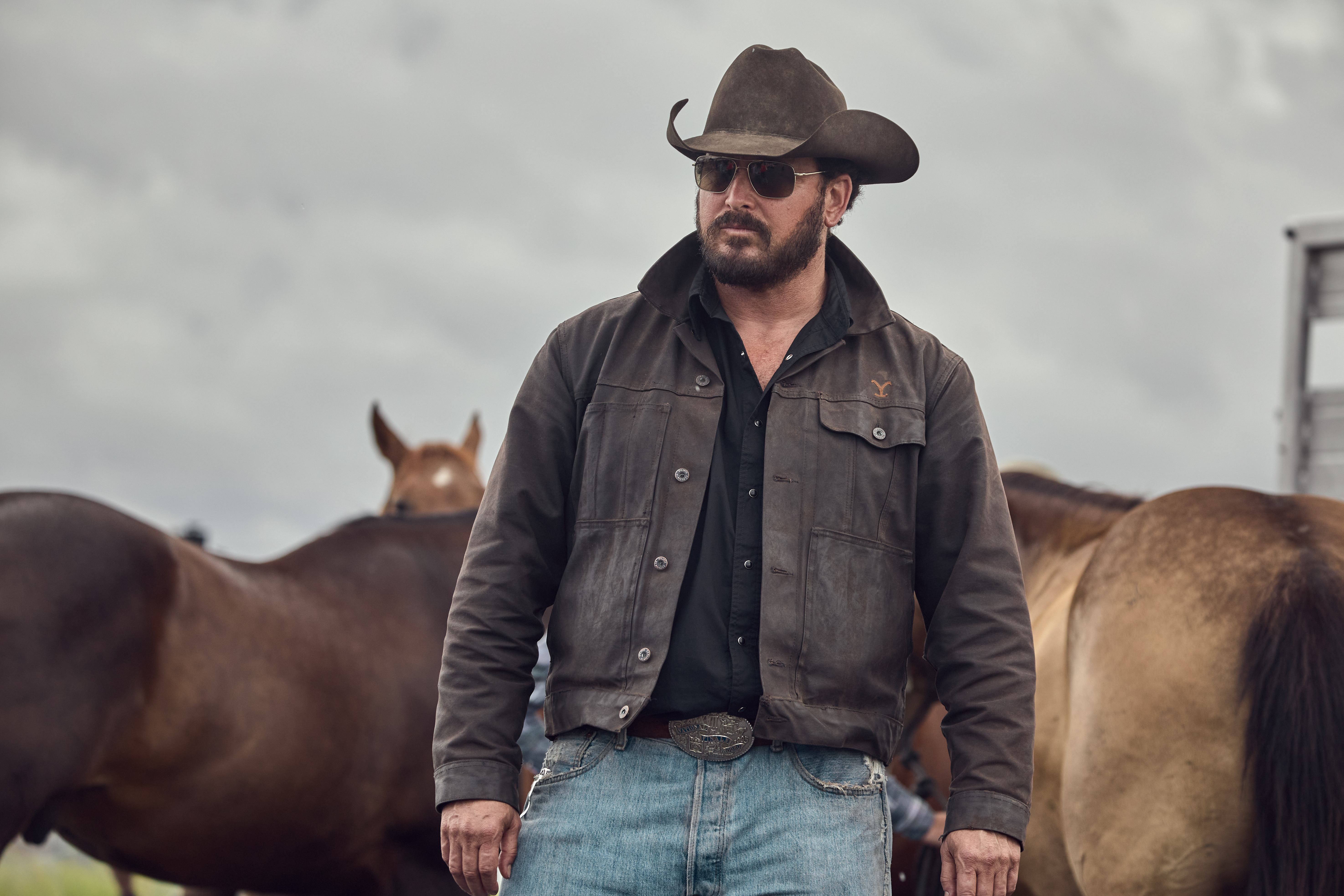 Rip Wheeler walks through a field of horses in a cowboy hat and shades.