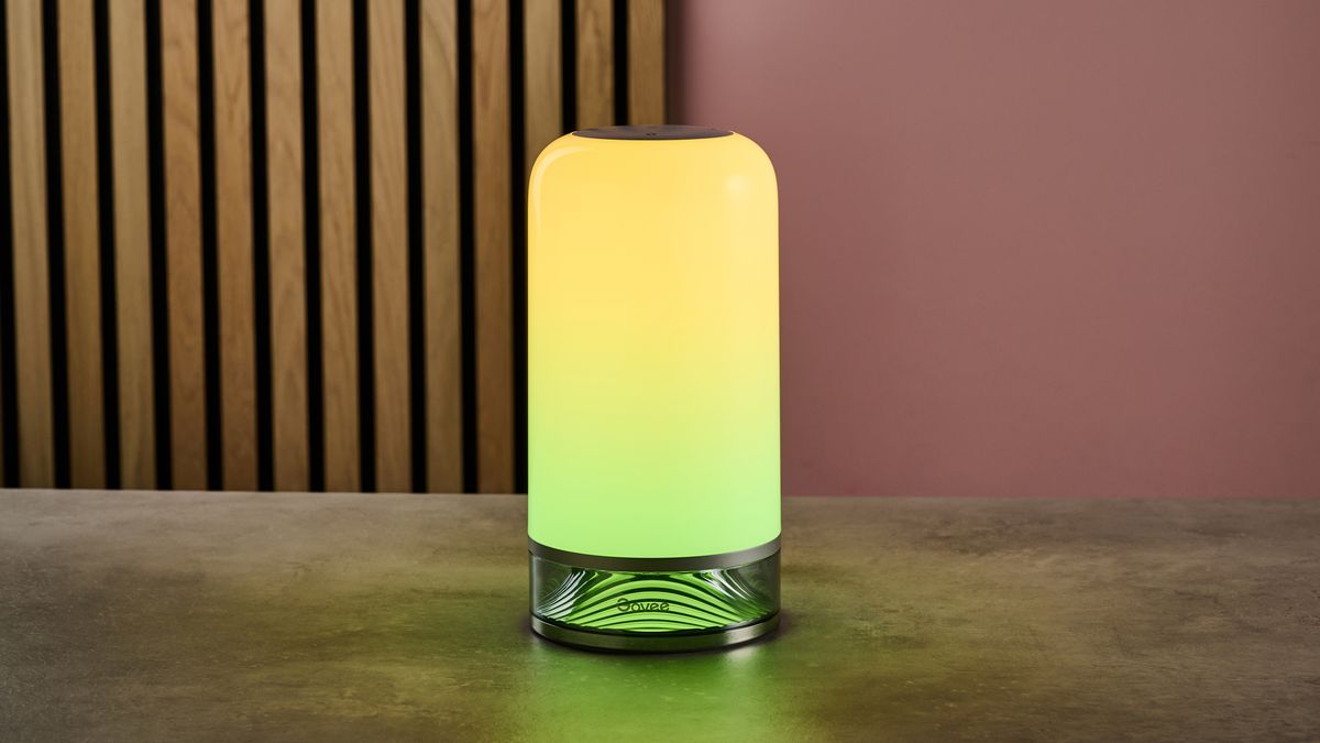 The Govee Table Lamp 2 sits on a stone-effect table in a dimly-lit pink room. It is lit up with yellow at the top, transitioning in a gradient to green at the base,