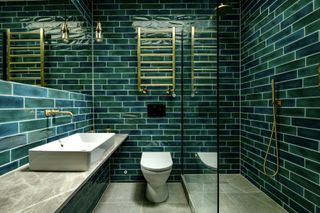 A green bathroom with green wall tiles and brass fixtures and fittings and a glass shower door