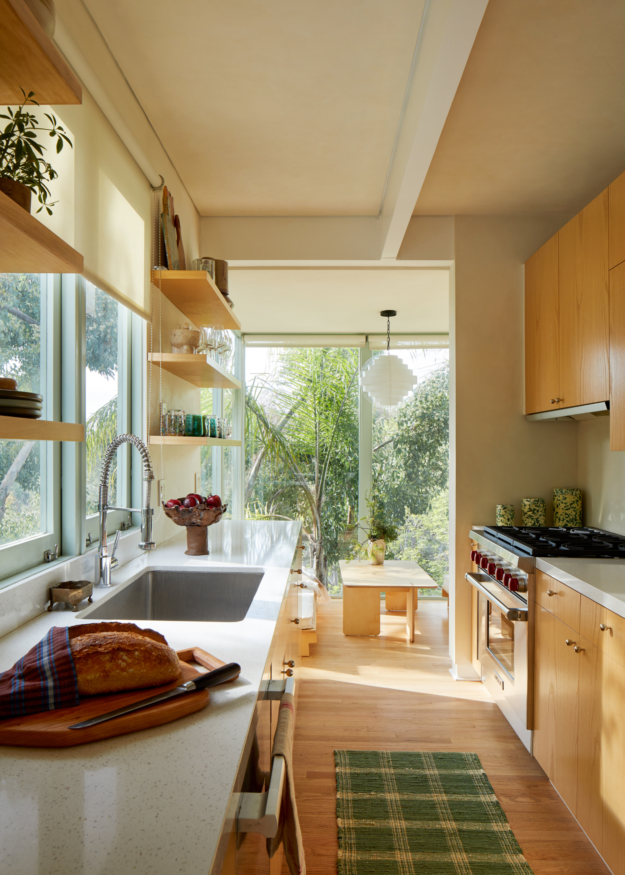 A bright kitchen with wood shelves and cabinets, a runner on the floor, a chopping board with a knife and a loaf of bread, a pedestal bowl of fruit and a soap dish