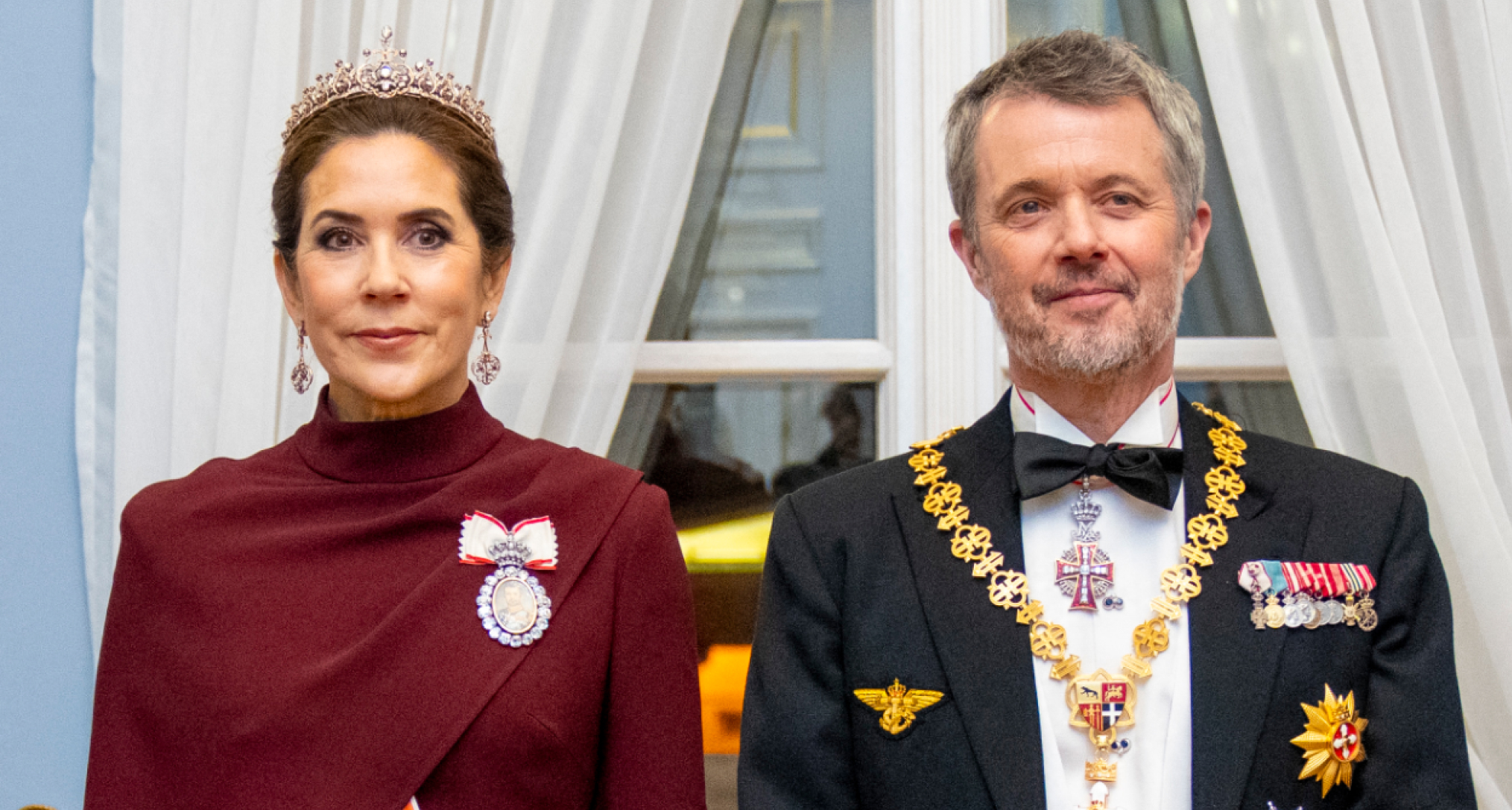 Queen Mary in a maroon gown and tiara standing with King Frederik in a military uniform and bow tie