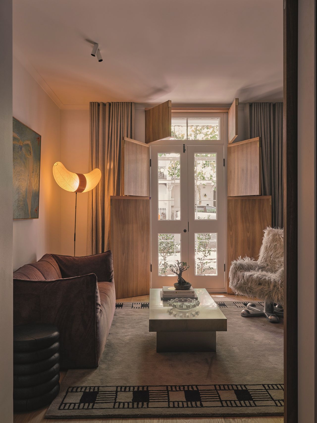 living room with grey curtains and timber shutters, a brown velvet sofa, shaggy armchair, rectangle coffee table on patterned rug