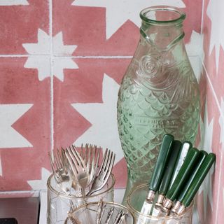 A fish-shaped green glass bottle with glasses of cutlery against a tiled kitchen splashback