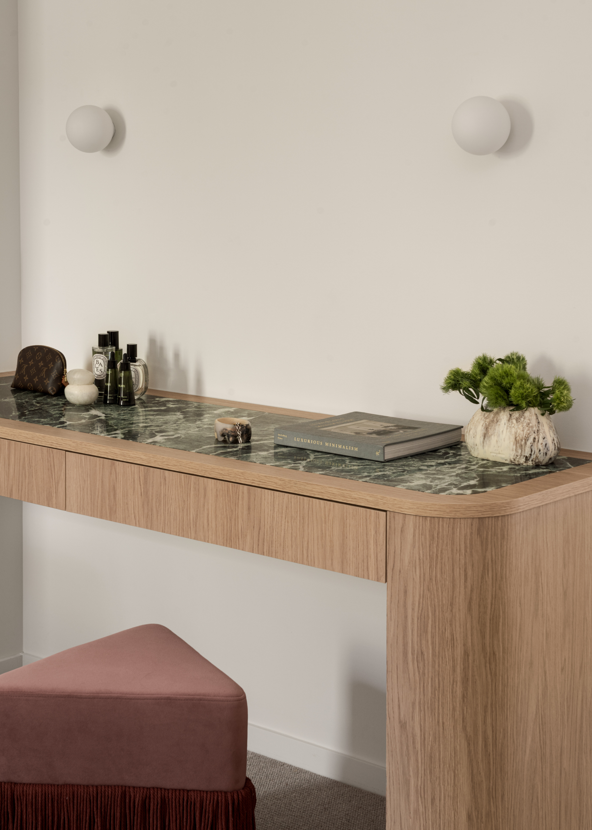 A vanity space with a fringe stool, a wood table with a green marble inlay. Also spotted are perfumes, skin care, a book, and a vase of stems